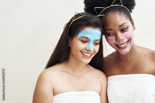 Multimasking. Two different ethnic girls are hugging. The girls' faces are covered with pink and blue face care masks. Beauty face, facial treatment and spa concept photo