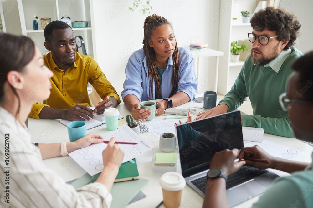 Portrait of contemporary multi-ethnic team discussing business project while sitting at cluttered table in conference room and listening to manager, copy space