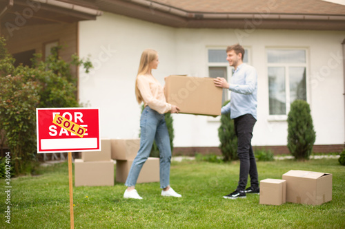 Millennial couple with carrying box in front of new home on moving day, focus on SOLD sign. Copy space