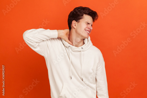 Unhappy worried young man in casual white sweatshirt with hood massaging numb neck, muscle tension, spine injury, osteochondrosis. Indoor studio shot isolated on orange background