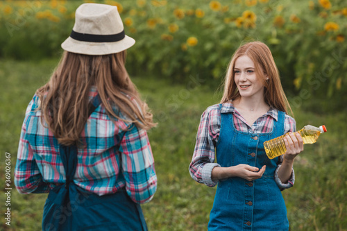 Two young farmerettes having a discussion while one of them is holding a bottle of golden sunflower oil in the middle of a green blooming sunflower field.