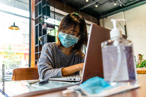 Young asian woman using laptop computer working at home with alcohol gel in quarantine for coronavirus wearing protective mask with social distancing and .