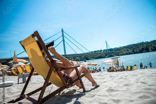 An attractive girl in a black swimsuit lies on a yellow wooden deck chair at a beach bar. The beach is sandy and behind you can see the bridge.