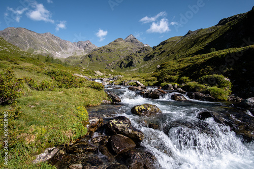 Bergbach (Debantbach bei Lienz) in Österreich mit Steinen und grünen Wiesen, im Hintergrund hohe Berge (Glödis, Debantgrad, Hochschober, Nationalpark hohe Tauern)