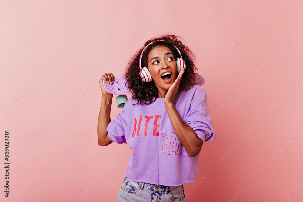 Blissful black girl with curly hairstyle posing with skateboard. Good-humoured african female model in casual clothes listening music. Blissful black girl with curly hairstyle posing with skateboard. Good-humoured african female model in casual clothes listening music.