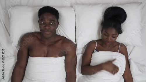 Close up portrait of young African couple lying in bed under blanket looking embarrassed before first sex. Girl turning to boyfriend and hinting about her desire to make love