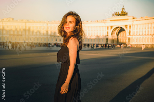 A young woman in a dress with long hair walks on Palace square at sunset in Saint Petersburg