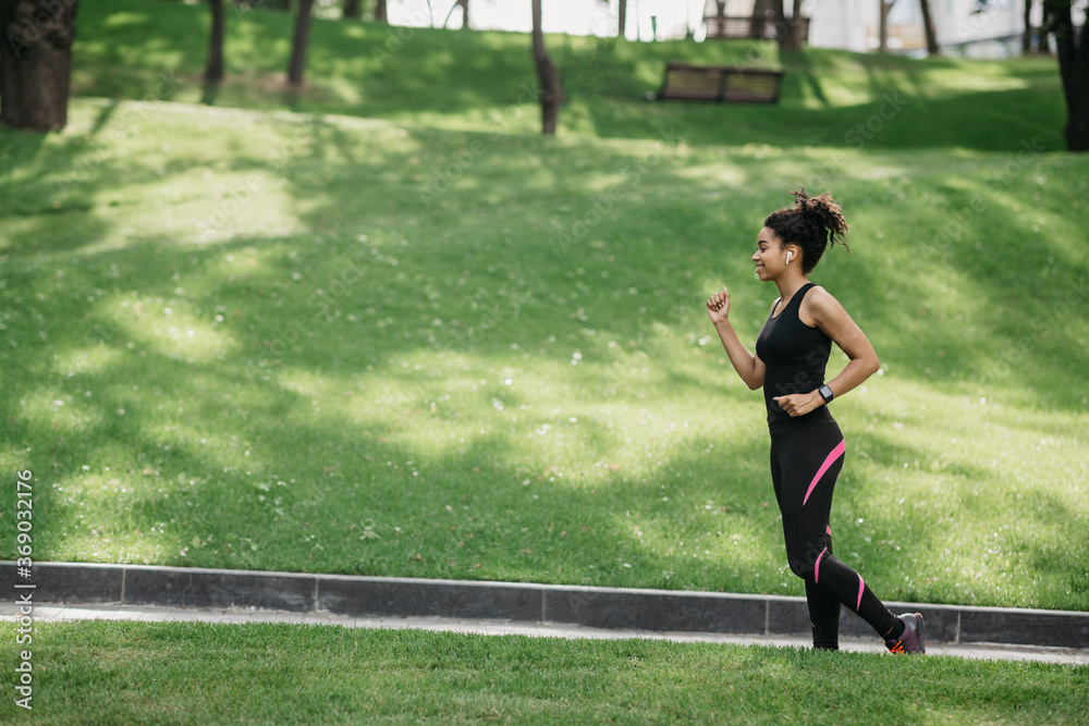 Jogging in park. Smiling fit african american woman with wireless headphones and fitness tracker jogging Jogging in park. Smiling fit african american woman with wireless headphones and fitness tracker jogging
