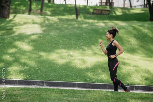 Jogging in park. Smiling fit african american woman with wireless headphones and fitness tracker jogging © Prostock-studio