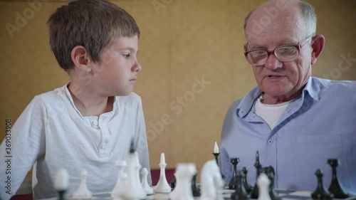 family recreation, simpatic joyful boy playing with his elderly grandfather in glasses for sight board game of chess sitting at table in room photo