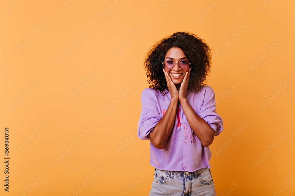 Pleased black girl in trendy sunglasses having fun during indoor photoshoot. Refined stylish african woman playfully smiling on yellow background.