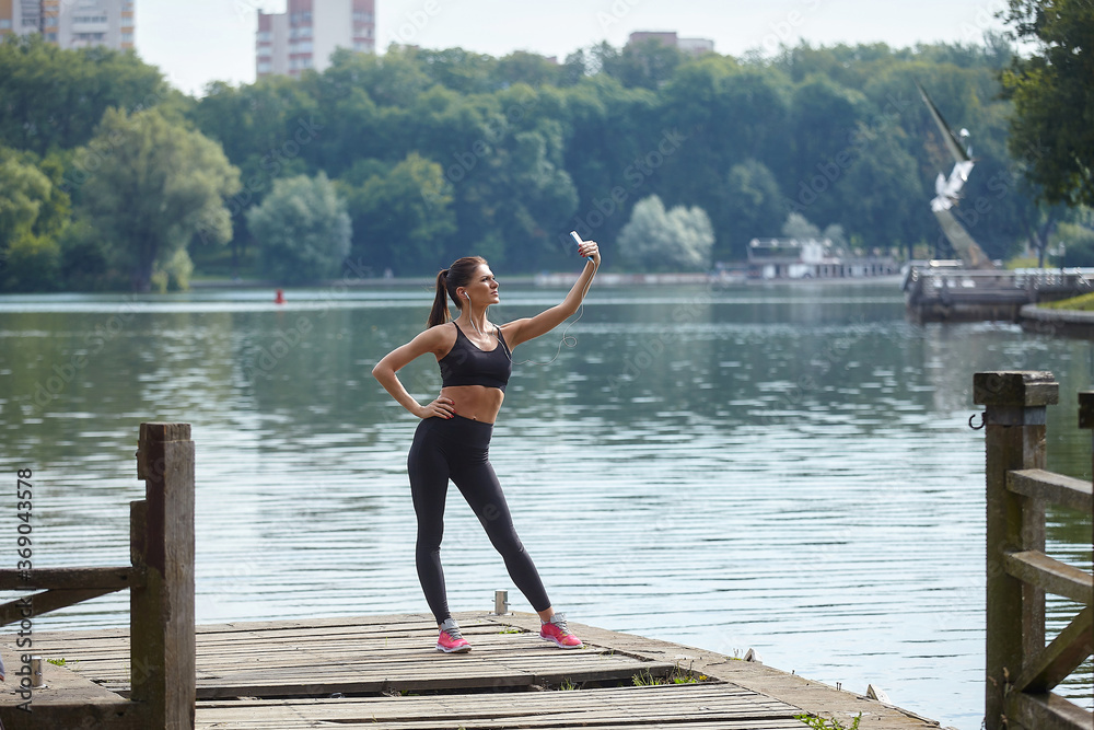 Distance learning concept. An attractive girl in sportswear stands on a wooden pier in a city park. Online lessons via smartphone.