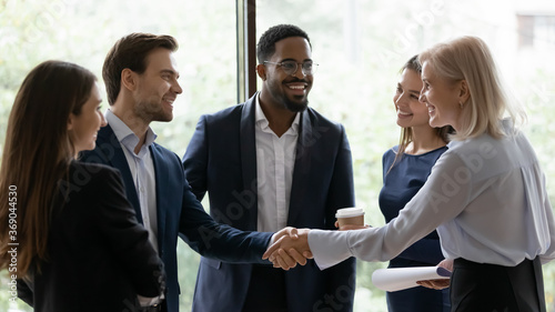 50s business lady shake hands greeting caucasian millennial businessman surrounded by multi ethnic businesspeople in formal suits. Sign contract express trust, partnership, start group meeting concept