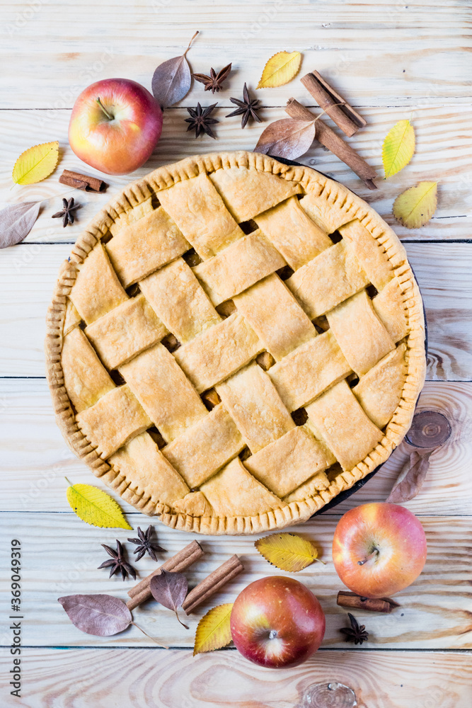 Homemade apple pie with ripe apples, species and autumn leaves on light rustic wooden background, top view