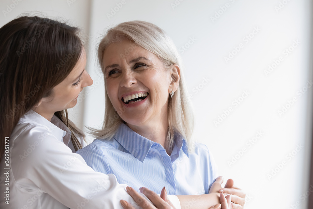 Loving adult daughter hugs cheerful elderly mother close up. Picture of happy grown up child cuddles mommy from behind, pretty women enjoy tender moment looking at each other. Love and warmth concept Loving adult daughter hugs cheerful elderly mother close up. Picture of happy grown up child cuddles mommy from behind, pretty women enjoy tender moment looking at each other. Love and warmth concept