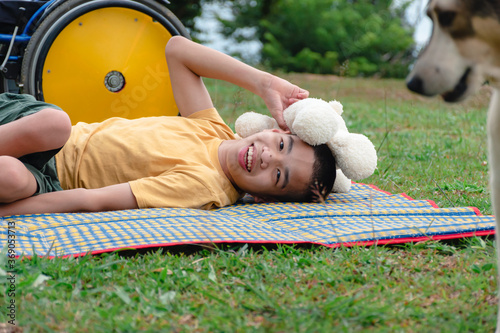 Asian special child playing on picnic mat happily with his pet near the wheelchair,Green nature background,Natural therapy,Life in the education age of disabled children, Happy disability kid concept.
