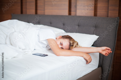 Young girl wake up with phone under blanket in white bed in bedroom. Healthy sleep, healthy sleep apps, lazy morning