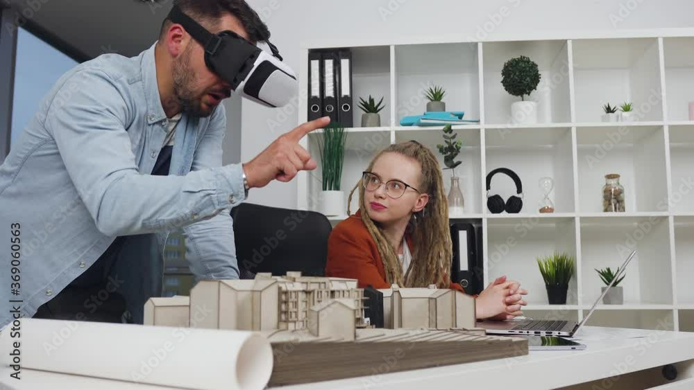 Handsome experienced purposeful young bearded architect in special 3d glasses talking with his stylish colleague with dreadlocks which entering project measurements of new buildings into computer Handsome experienced purposeful young bearded architect in special 3d glasses talking with his stylish colleague with dreadlocks which entering project measurements of new buildings into computer