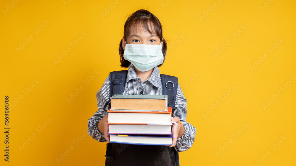 Thai young girl wearing face mask, Asian kid holding lots of books isolated on yellow or orange background, learning and back to school during coronavirus or covid 19 - obrazy, fototapety, plakaty 