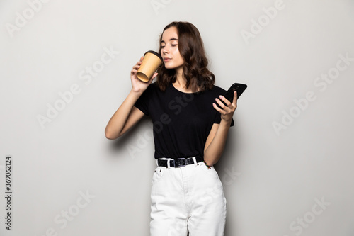 Photo of successful woman in formal wear standing with smartphone and takeaway coffee in hands over gray background