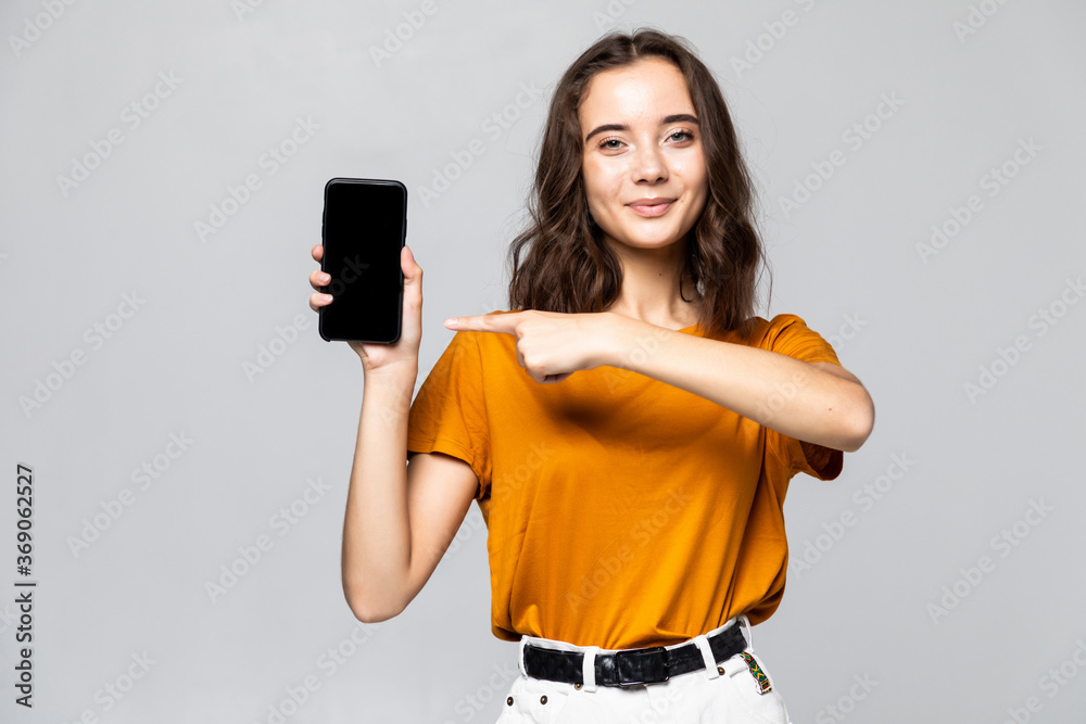 © dianagrytsku - Happy woman in casual clothes showing blank smartphone screen and looking at the camera over grey background