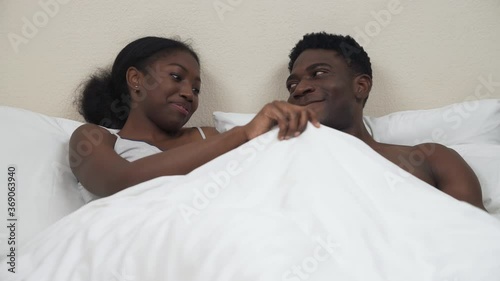 Portrait of African American couple lying in bed. Woman lifting blanket and expressing amazement because of man's penile erection. Sexual life concept