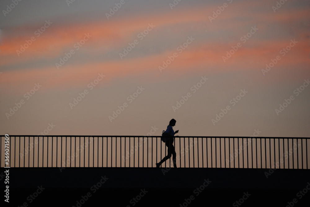 Silhouette of a woman with a backpack looking at her cell phone crossing a bridge at sunset