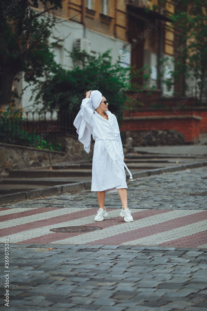 A glamorous lady in a white bathrobe with a white towel on her head and elegant sunglasses is walking along the street of a big city