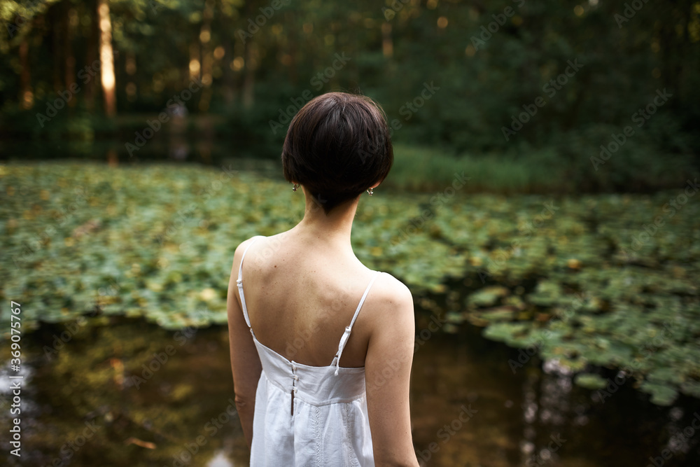 Rear shot of unrecognizable short haired young woman in strap white dress relaxing by pond in park, enjoying beautiful landscape and hot summer day. Back view of female walking outdoors alone Rear shot of unrecognizable short haired young woman in strap white dress relaxing by pond in park, enjoying beautiful landscape and hot summer day. Back view of female walking outdoors alone