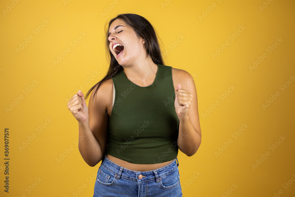 Young pretty woman over yellow background very happy and excited making winner gesture with raised arms, smiling and screaming for success. Young pretty woman over yellow background very happy and excited making winner gesture with raised arms, smiling and screaming for success.