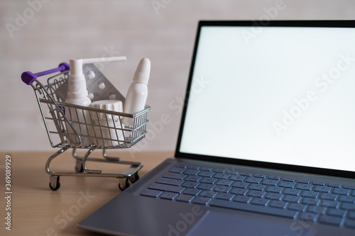 Laptop with blank white screen and miniature shopping cart full of medicines. Online pharmacy concept. Website for buying tablets.