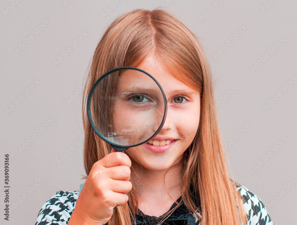 Child girl student looking through magnifying glass on gray background with available copy space. Back to school concept. Child girl student looking through magnifying glass on gray background with available copy space. Back to school concept.