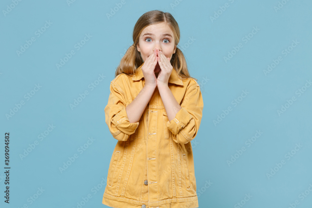 Surprised little blonde kid girl 12-13 years old in yellow jacket posing isolated on pastel blue background studio portrait. Childhood lifestyle concept. Mock up copy space. Covering mouth with hands.