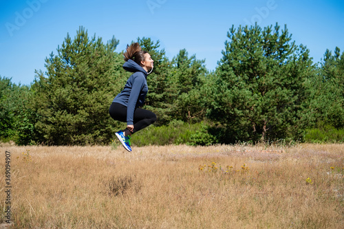 Young woman in sports outfit hops high on a natural meadow with trees and blue sky in the background