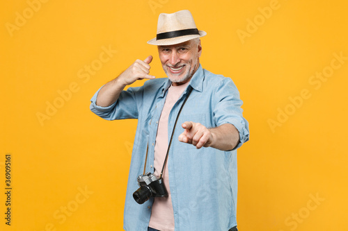 Smiling traveler tourist elderly gray-haired man in hat isolated on yellow background. Passenger traveling abroad on weekends. Doing phone gesture like says call me back  point index finger on camera.
