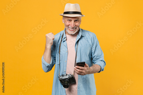 Happy traveler tourist elderly gray-haired man isolated on yellow background. Passenger traveling abroad on weekends. Air flight journey. Using mobile phone booking taxi hotel doing winner gesture.