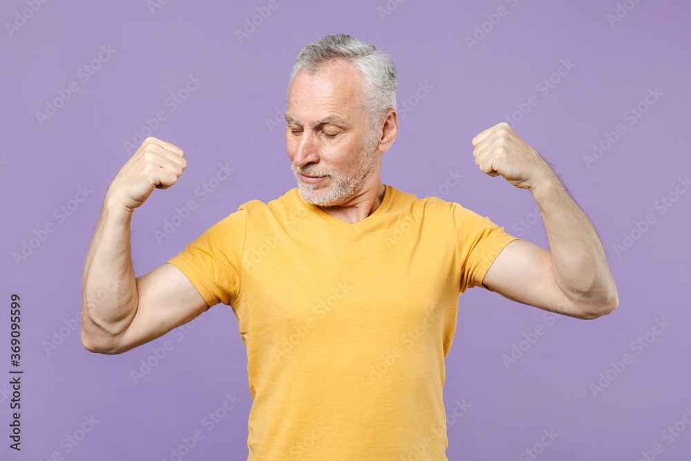 Strong elderly gray-haired mustache bearded man in casual yellow t-shirt posing isolated on pastel violet wall background studio. People lifestyle concept. Mock up copy space. Showing biceps, muscles. Strong elderly gray-haired mustache bearded man in casual yellow t-shirt posing isolated on pastel violet wall background studio. People lifestyle concept. Mock up copy space. Showing biceps, muscles.