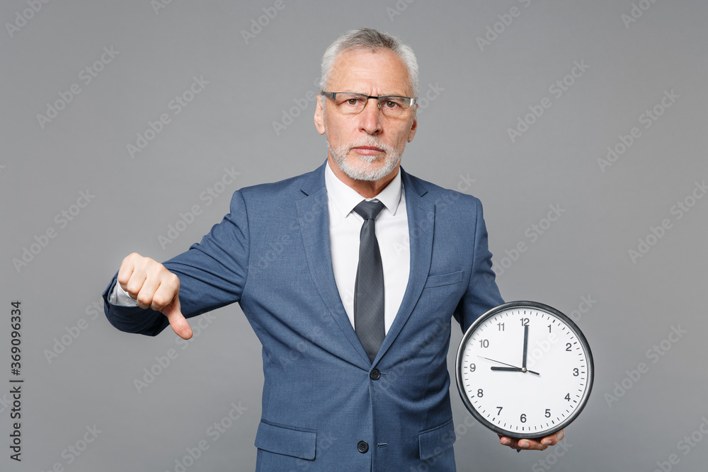 Displeased elderly gray-haired bearded business man in blue suit shirt tie isolated on grey wall background studio portrait. Achievement career wealth business concept. Hold clock, showing thumb down. Displeased elderly gray-haired bearded business man in blue suit shirt tie isolated on grey wall background studio portrait. Achievement career wealth business concept. Hold clock, showing thumb down.