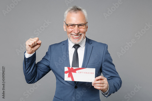 Joyful elderly gray-haired business man in classic blue suit shirt isolated on grey background studio portrait. Achievement career wealth business concept. Hold gift certificate doing winner gesture.