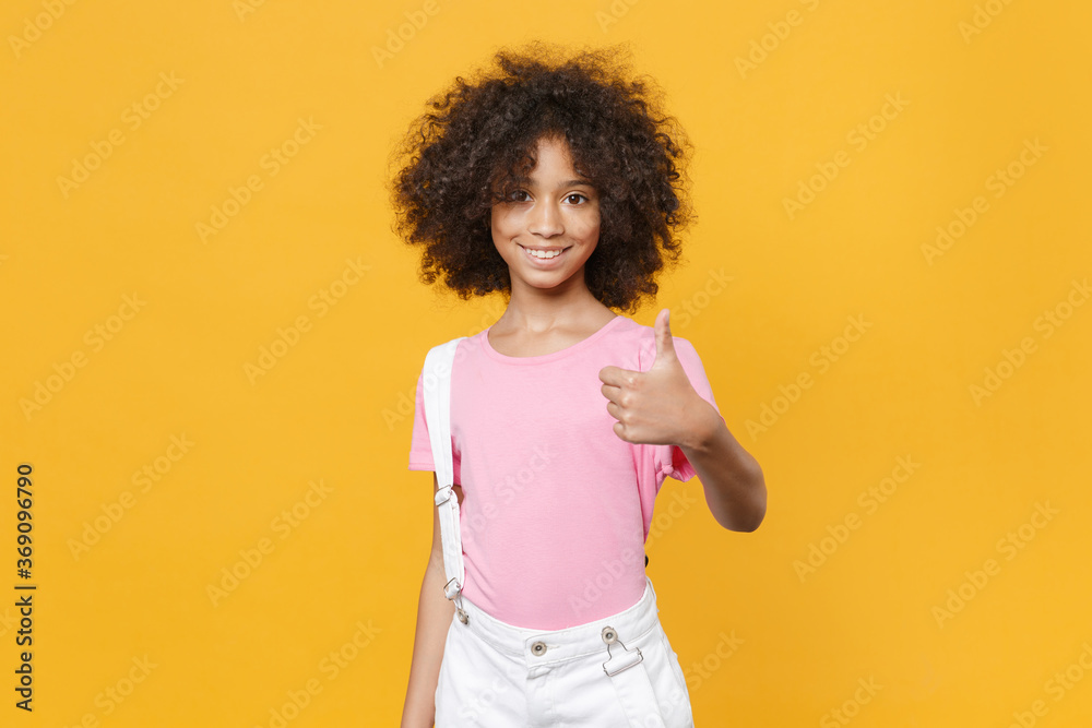 Smiling little african american kid girl 12-13 years old in pink t-shirt isolated on yellow wall background children studio portrait. Childhood lifestyle concept. Mock up copy space. Showing thumb up. Smiling little african american kid girl 12-13 years old in pink t-shirt isolated on yellow wall background children studio portrait. Childhood lifestyle concept. Mock up copy space. Showing thumb up.
