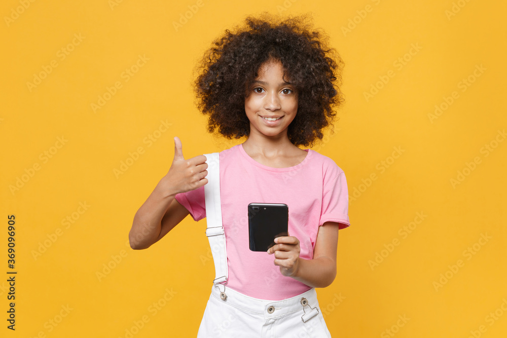 Smiling little african american kid girl 12-13 years old in pink t-shirt isolated on yellow wall background studio. Childhood lifestyleconcept. Mock up copy space. Using mobile phone showing thumb up. Smiling little african american kid girl 12-13 years old in pink t-shirt isolated on yellow wall background studio. Childhood lifestyleconcept. Mock up copy space. Using mobile phone showing thumb up.