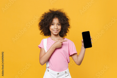 Smiling african american kid girl 12-13 years old in pink t-shirt isolated on yellow background. Childhood concept. Mock up copy space. Pointing index finger on mobile phone with blank empty screen.
