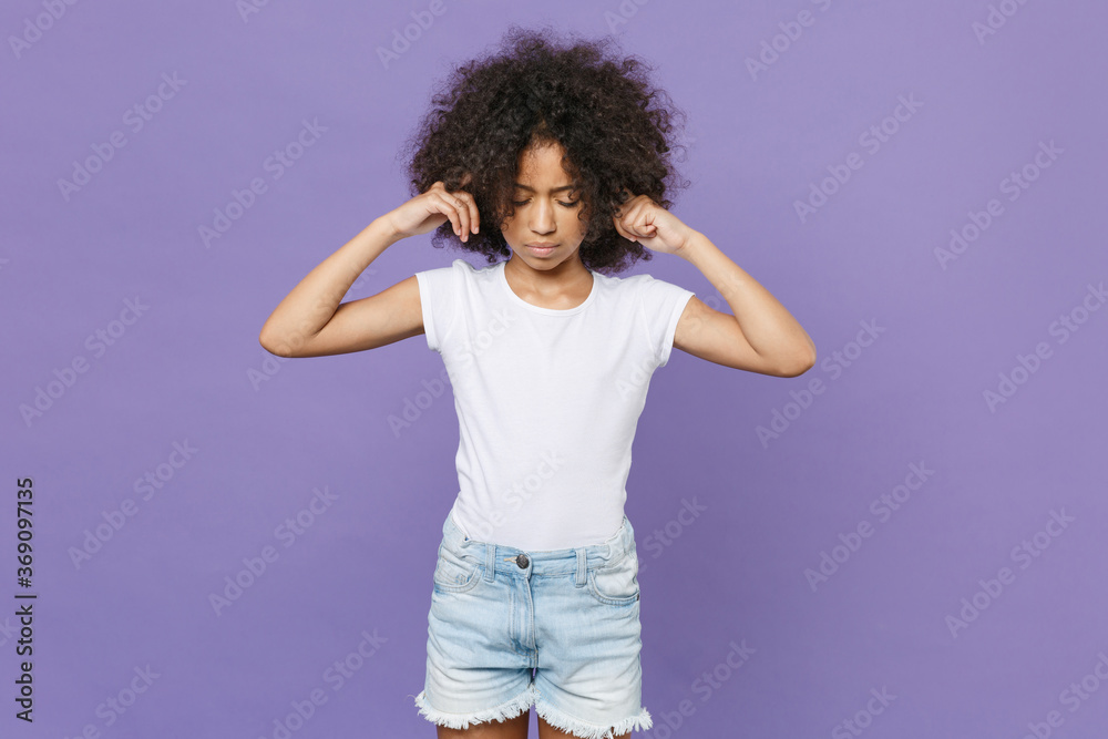 Displeased little african american kid girl 12-13 years old in white t-shirt isolated on violet background studio portrait. Childhood lifestyle concept. Mock up copy space. Covering ears with fingers. Displeased little african american kid girl 12-13 years old in white t-shirt isolated on violet background studio portrait. Childhood lifestyle concept. Mock up copy space. Covering ears with fingers.