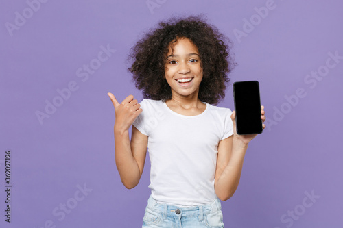 Smiling little african american kid girl 12-13 years old in white t-shirt isolated on violet wall background. Childhood lifestyle concept. Hold mobile phone with blank empty screen, showing thumb up.