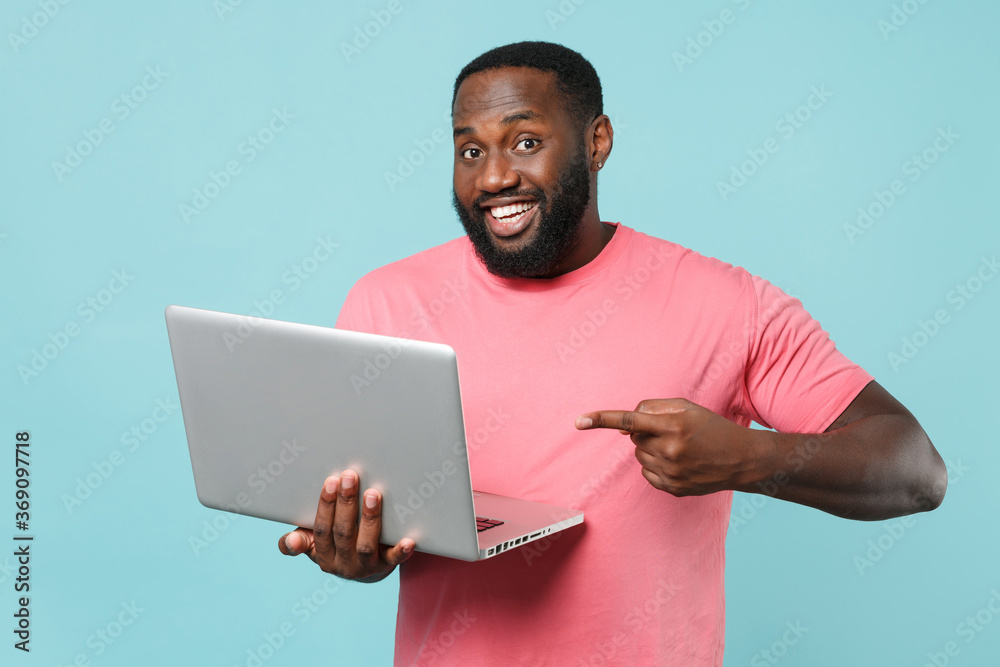 Smiling young african american man guy in casual pink t-shirt isolated on blue background studio portrait. People lifestyle concept. Mock up copy space. Pointing index finger on laptop pc computer. Smiling young african american man guy in casual pink t-shirt isolated on blue background studio portrait. People lifestyle concept. Mock up copy space. Pointing index finger on laptop pc computer.