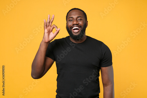 Cheerful african american man guy football fan in casual black t-shirt isolated on yellow wall background studio portrait. People emotions lifestyle concept. Mock up copy space. Showing OK gesture.
