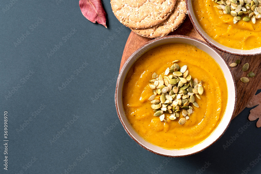 Pumpkin soup with pumpkin seeds in bowls and dry autumn leaves on a dark gray background top view. Free space for text. Copy space. Flat lay. Autumn food. American cuisine. Pumpkin soup with pumpkin seeds in bowls and dry autumn leaves on a dark gray background top view. Free space for text. Copy space. Flat lay. Autumn food. American cuisine.
