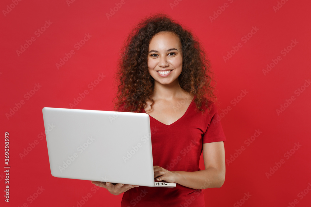 Smiling young african american woman girl in casual t-shirt posing isolated on red background studio portrait. People emotions lifestyle concept. Mock up copy space. Working on laptop pc computer. Smiling young african american woman girl in casual t-shirt posing isolated on red background studio portrait. People emotions lifestyle concept. Mock up copy space. Working on laptop pc computer.