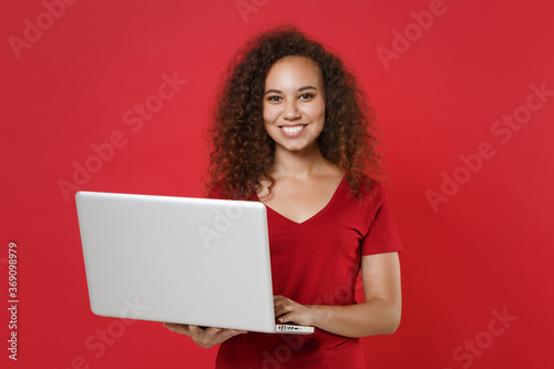 Smiling young african american woman girl in casual t-shirt posing isolated on red background studio portrait. People emotions lifestyle concept. Mock up copy space. Working on laptop pc computer. © ViDi Studio