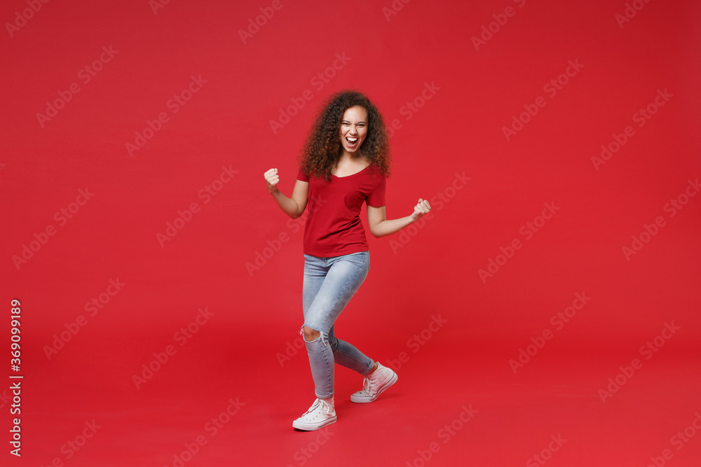 Full length portrait of happy young african american girl in casual t-shirt isolated on red background studio portrait. People emotions lifestyle concept. Mock up copy space. Doing winner gesture.